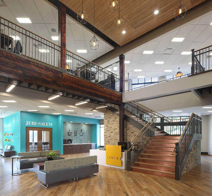 Multi-level Bader office lobby with grand staircase, reception desk, teal 'Jerusalem' wall, coffee bar, and lounge seating.