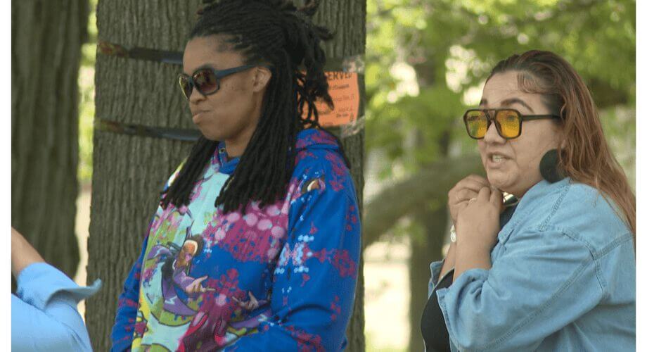 Two diverse women outdoors. One with dreadlocks and a colorful hoodie, the other in a denim jacket with yellow sunglasses.