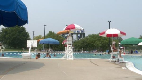 Lively outdoor aquatic center with swimmers, tall water slide, colorful umbrellas, and attentive lifeguards during summer.