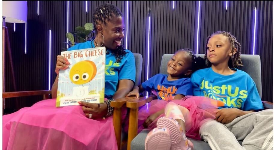 Mother and two kids, a girl and a boy, reading "The Big Cheese" book together. They are wearing JustUs Books t-shirts.