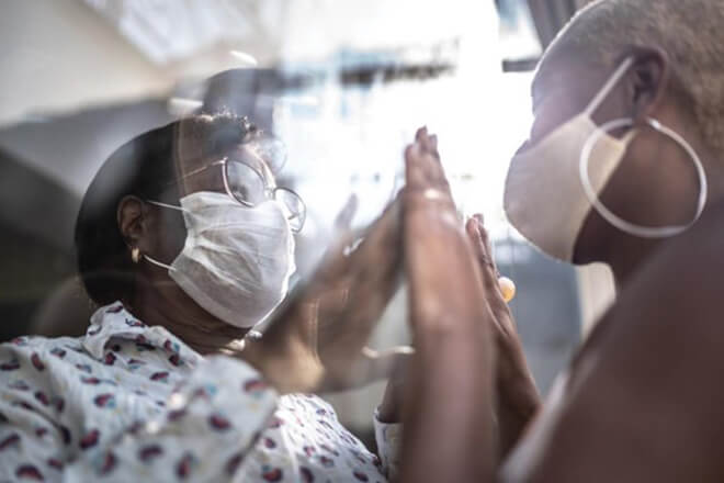 Two masked individuals press hands together through a glass barrier, symbolizing connection and love during social distancing and safe visits.