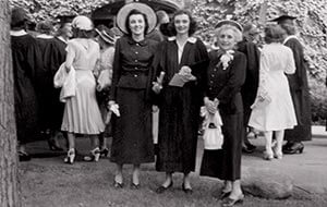 Black & white photo of three women in formal attire and academic gowns at a vintage graduation event.