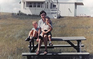 Mid-century family portrait with a mother and her two boys on a picnic bench on a sunny, grassy slope.