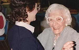 Happy senior woman with distinctive white glasses smiling, dancing with a younger woman. Joyful candid moment.