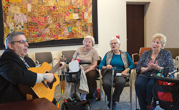Older man playing acoustic guitar for three senior women joyfully singing along in a senior living community.