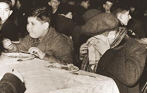 Vintage photo of boys eating a meal at an institution; one child covers his face, showing distress or sadness.