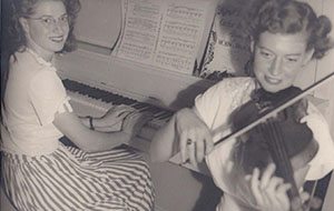 Vintage 1949 photo: Isabel playing piano, friend plays violin. Two smiling women musicians creating music.