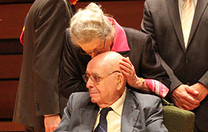 Isabel, an older woman, gently kisses an older man's forehead at a 2014 event, showing warmth and respect.
