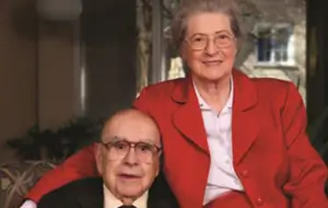 Elderly Isabel in red suit with her husband in dark suit, smiling for a portrait in 2018.