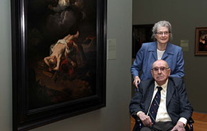 Isabel and partner, an elderly couple, visit an art museum in 2020, standing beside a large, dark classical painting.