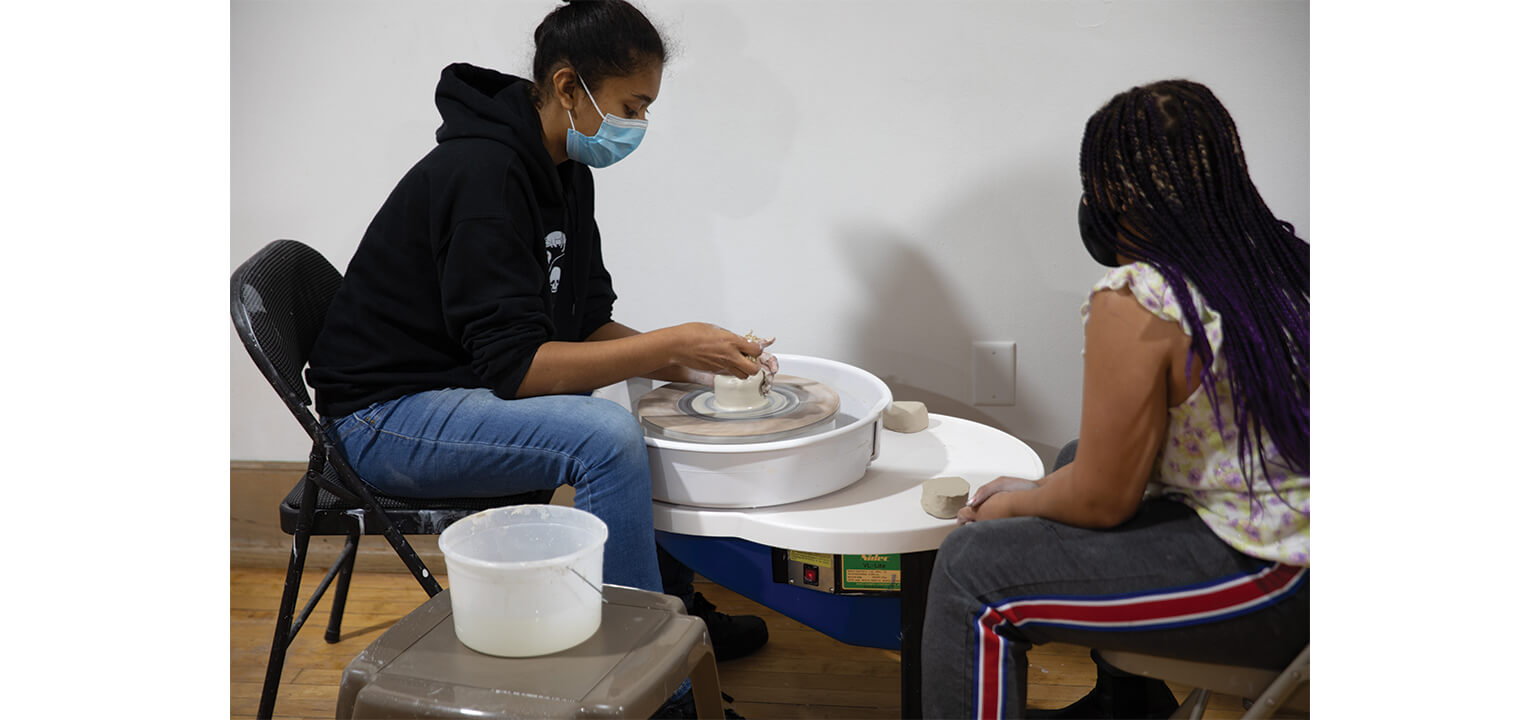 Masked adult guides student shaping clay on a pottery wheel. Hands-on art class, learning ceramics skills.