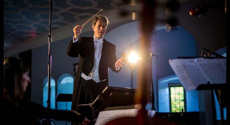 Male maestro in tuxedo conducting orchestra with baton during a live classical music concert.