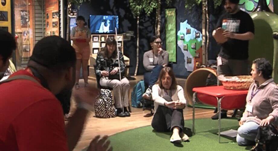 Adults engaging in a workshop within an interactive museum exhibit, featuring a presenter, artificial trees, and a world map.