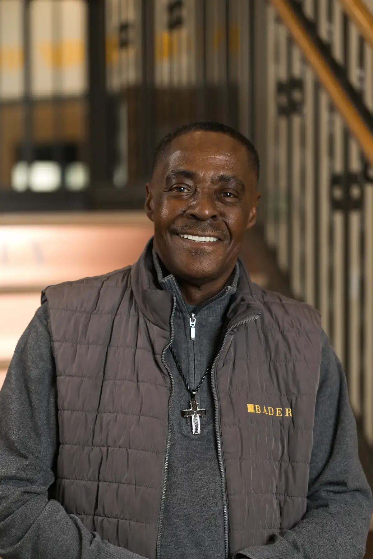 Smiling African American man wearing a grey puffer vest with the BADER logo and a cross pendant, professional portrait.