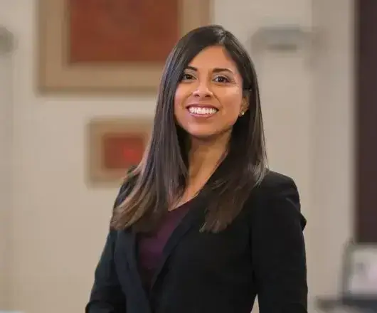 Smiling professional Latina businesswoman in black blazer. Confident corporate headshot, long dark hair, office setting.