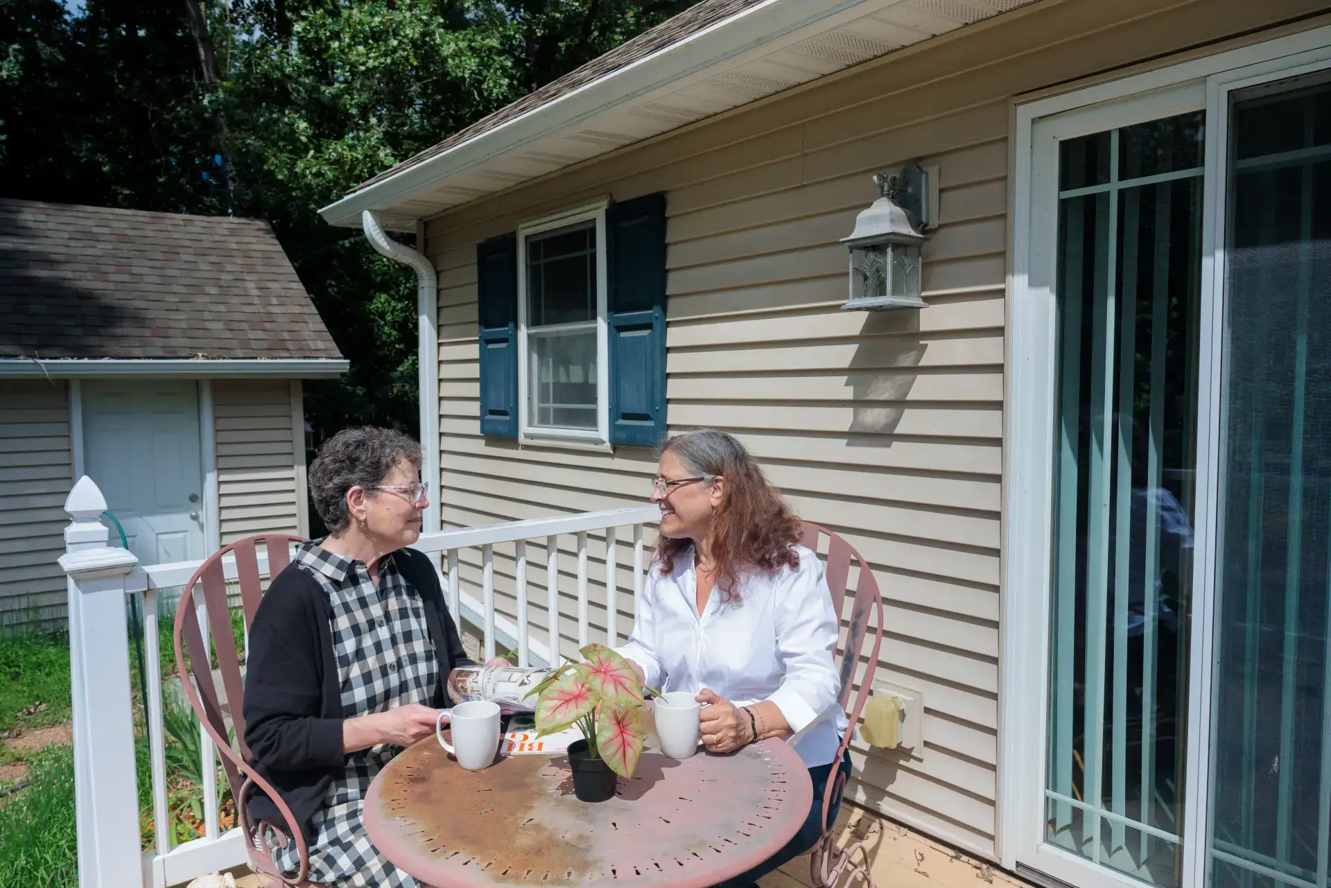 Two older women share a happy conversation on a sunny residential porch, enjoying coffee at The Comfort House.
