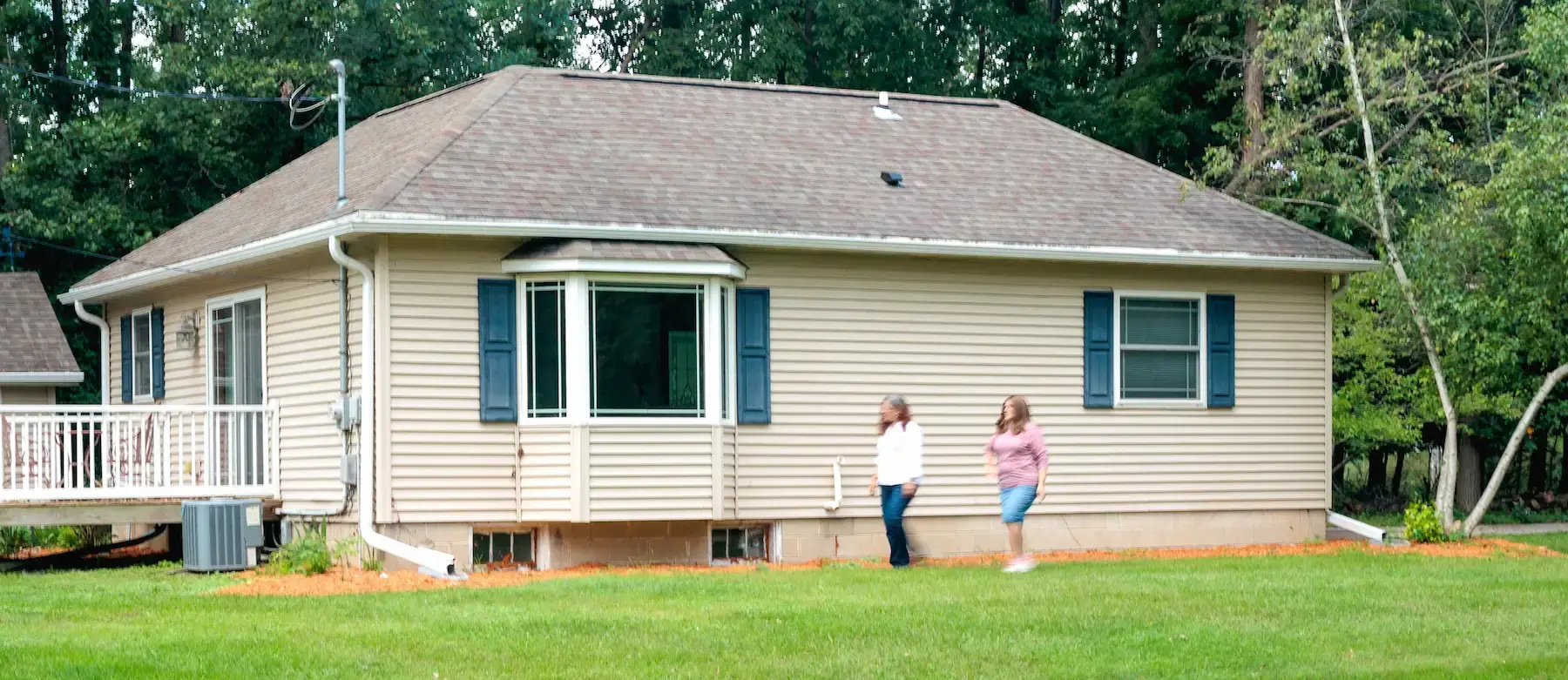 Beige residential house with bay window, blue shutters, white deck, two women on lawn, representing a Program Related Investment.