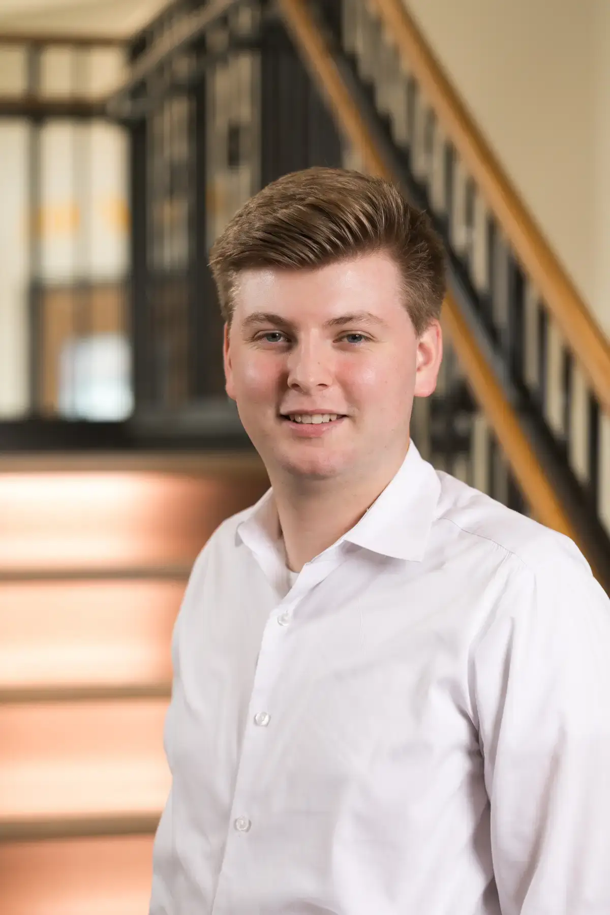 Professional headshot of Sam Smulyan smiling in a white collared shirt, with a modern office staircase background.