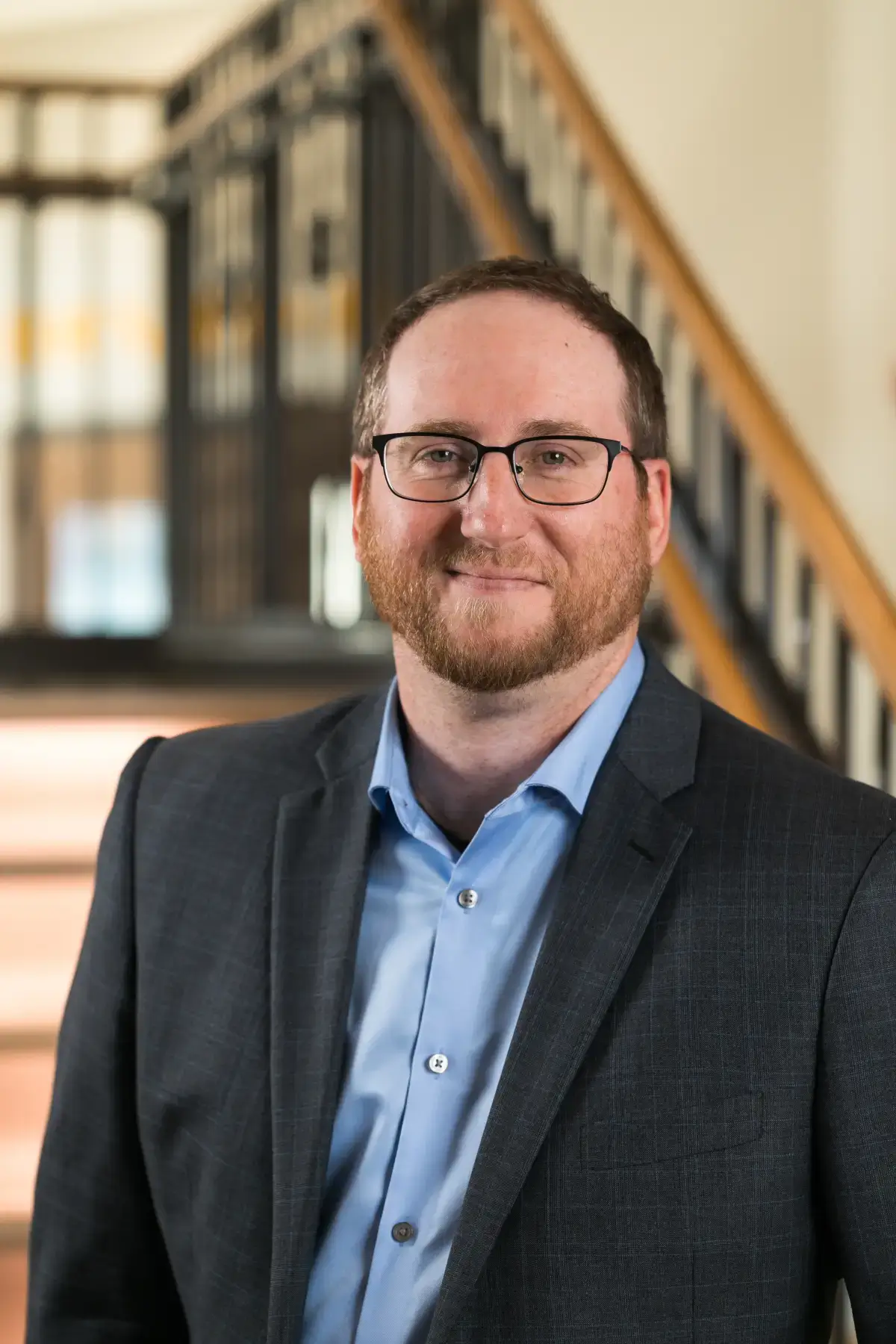 Tyler Pouros, smiling professional with glasses and beard, wearing a charcoal suit and light blue shirt in a modern office setting.
