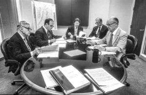 1994 corporate meeting with five men in suits around a round table, reviewing documents. Professional discussion.