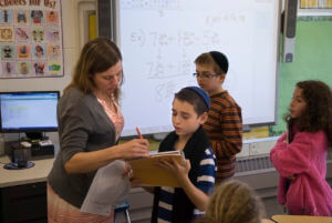 Teacher assists male student with math in a 2009 elementary classroom; other students and whiteboard with equations visible.