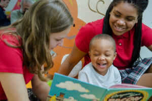 Two women and a happy toddler boy laughing during story time, engaged in early literacy and learning.