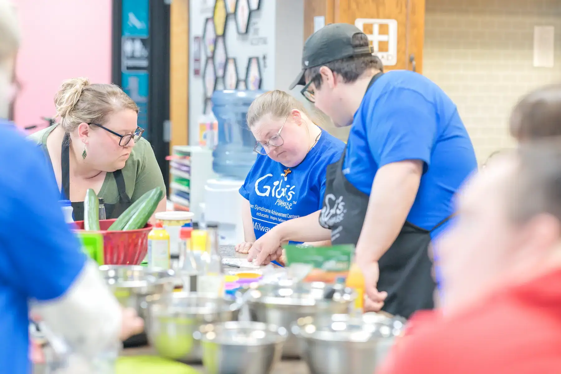 Participants in a Gigi's Playhouse cooking and life skills program, preparing fresh food like cucumbers in a kitchen.