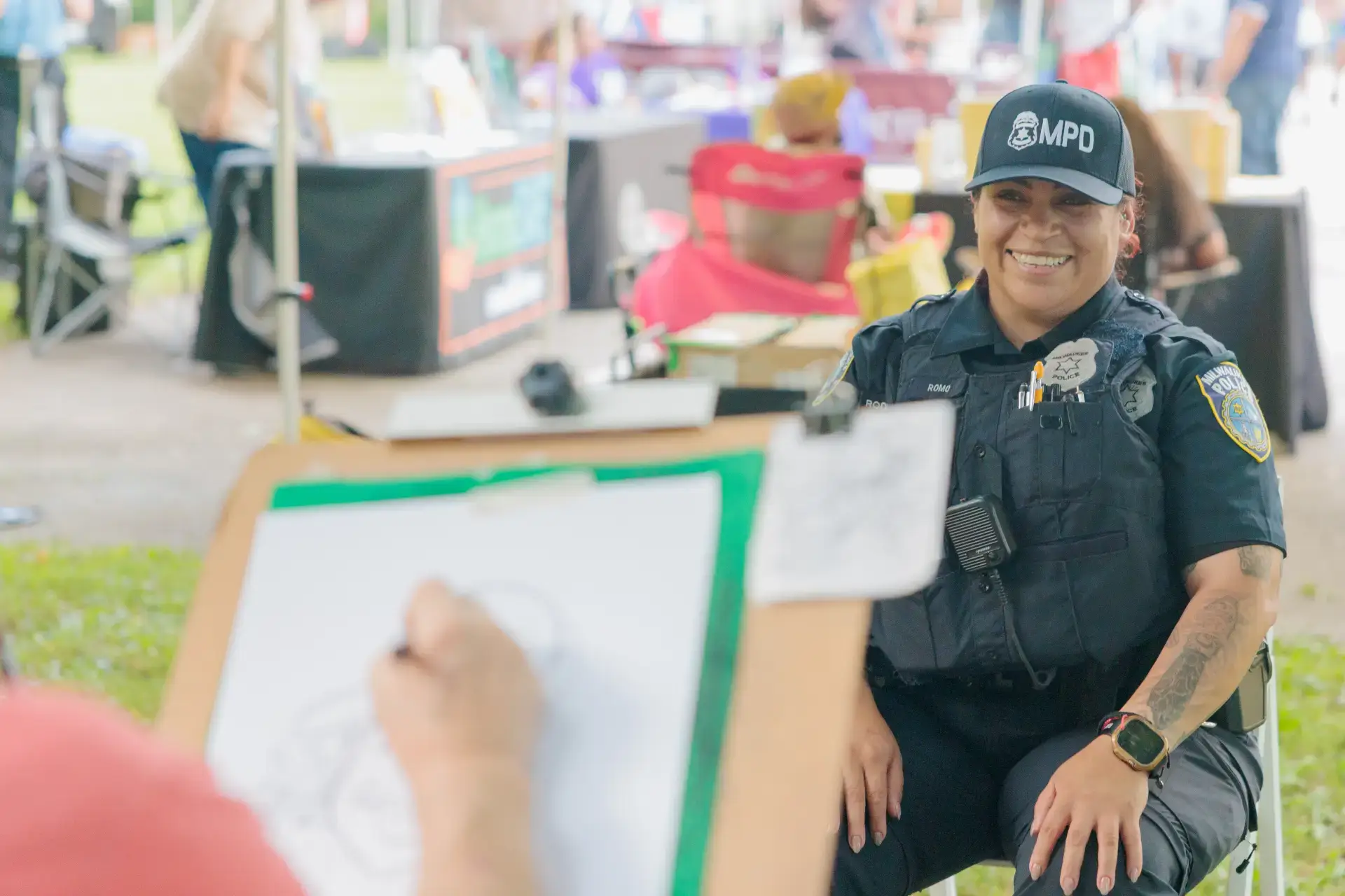 Smiling Milwaukee police officer (MPD) sitting at an outdoor community event while someone draws her portrait.