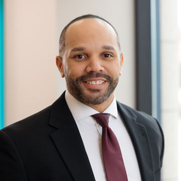 Marc Young smiling in a light grey suit, white shirt, and black and gold striped tie.