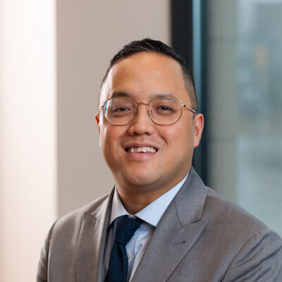 Marc Young smiling in a light grey suit, white shirt, and black and gold striped tie.