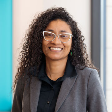 Yaa Engel smiling in glasses with long dark hair, blue shirt, and distinctive wooden and blue feathered earrings.